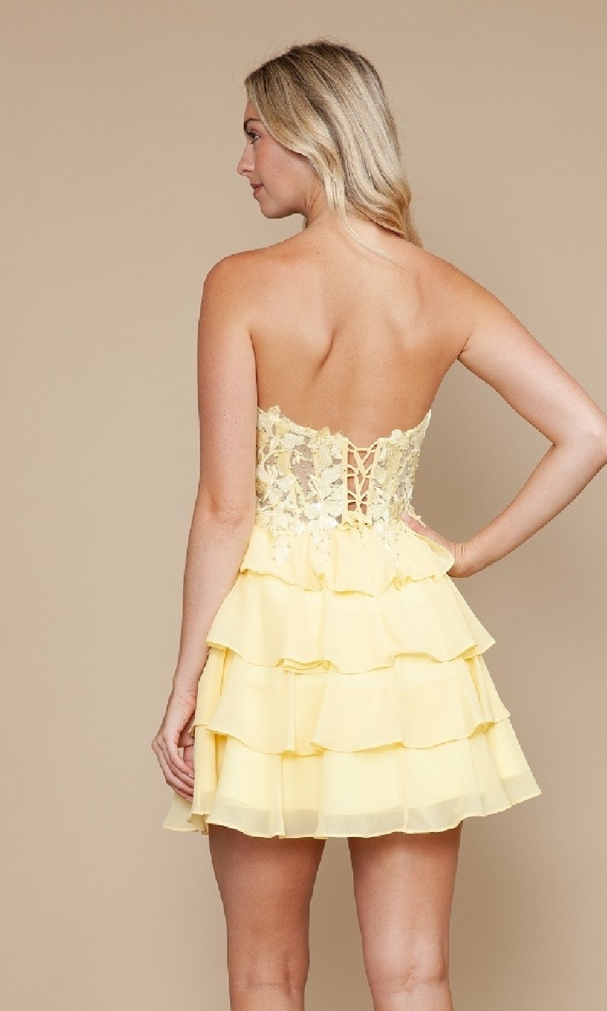 A woman faces away, modeling the Strapless Short Ruffled Hoco Dress: Poly USA 9838, which features a corset-style bodice and tiered ruffle skirt against a plain beige background.